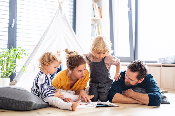 Young family with two small children indoors in bedroom reading a book.